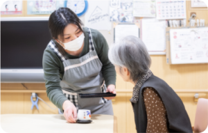 職員が高齢者と手工芸活動を行う様子。リハビリやレクリエーション場面。