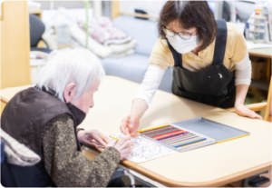 スタッフが高齢者と一緒に色鉛筆で描く活動。介護施設でのレクリエーション風景。